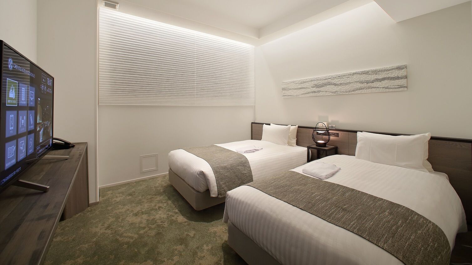 Une chambre d’hôtel avec deux lits, une télévision, une table de chevet, une lampe et une fenêtre avec des stores.