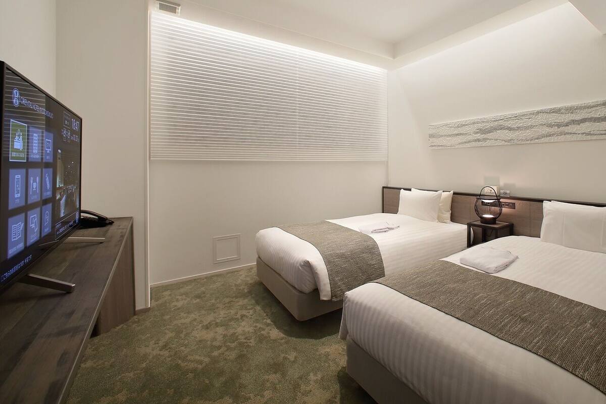 A hotel room with two beds, a TV, a bedside table, a lamp, and a window with blinds.