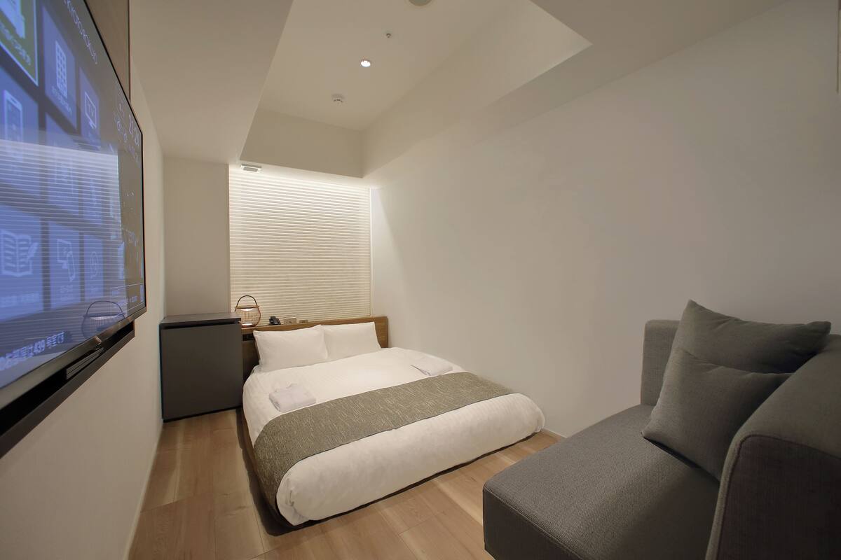 A modern hotel room with a bed, a sofa, a TV screen, and a window with blinds.