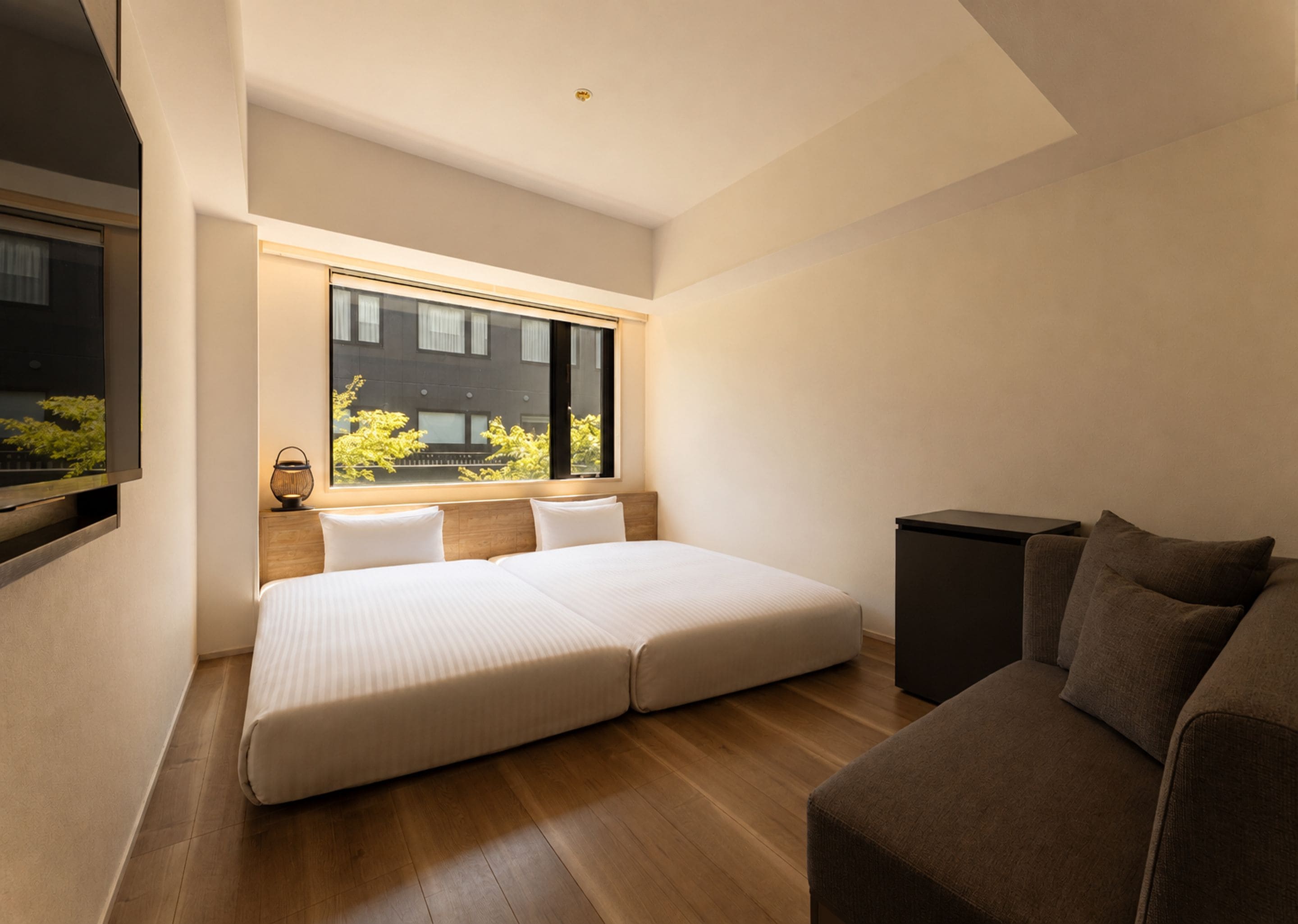 A modern bedroom with a large bed, a sofa, a TV, and a window with a view of greenery.