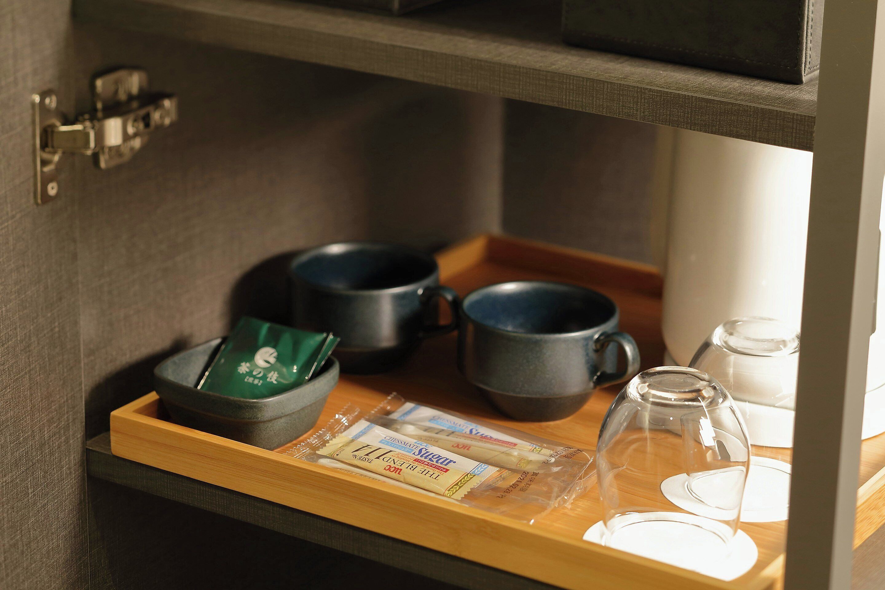 Un plateau en bois avec un sachet de thé, deux tasses foncées et un pichet en verre, le tout posé sur une étagère.