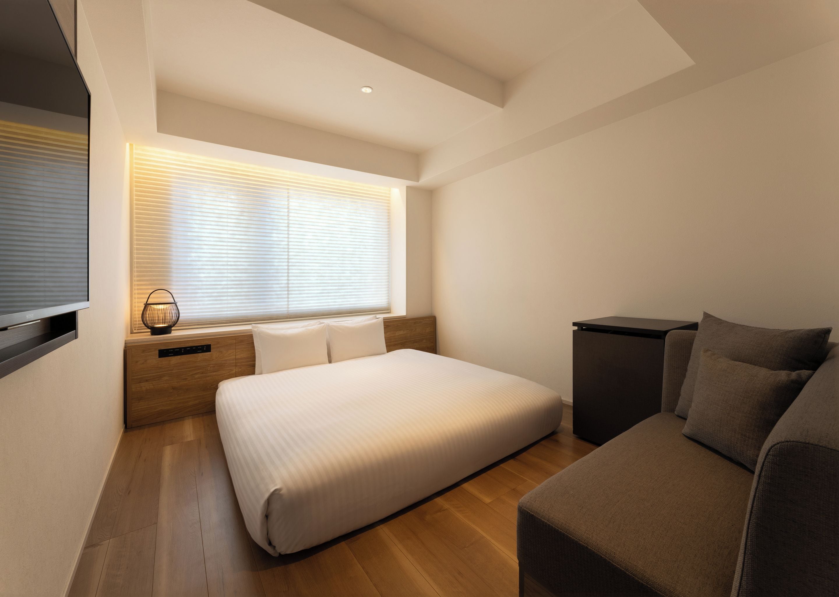 A modern bedroom with a bed, a sofa, a TV, and a window with blinds.