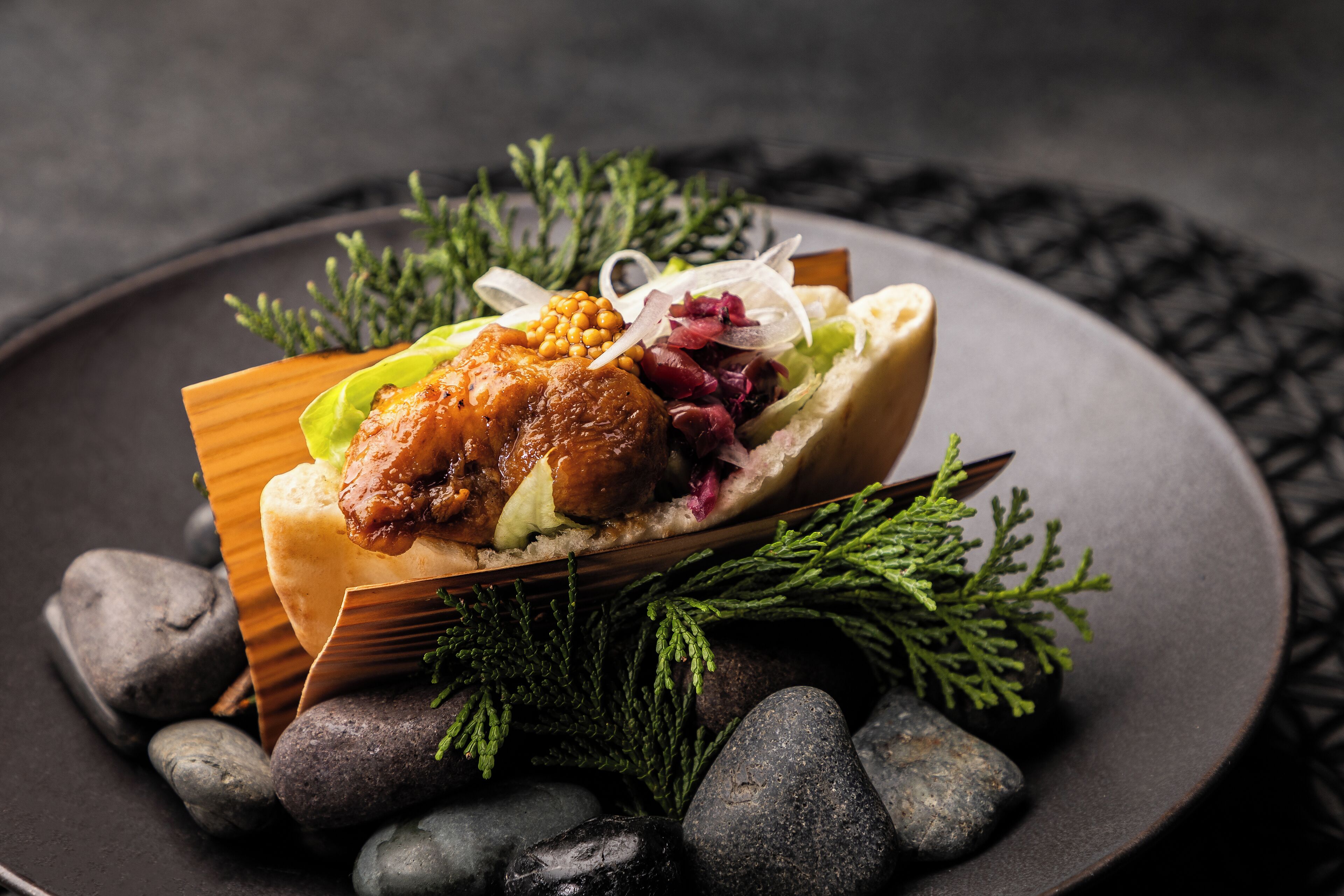 Un plato con carne, verduras y un pan sobre una vajilla oscura con detalles decorativos.