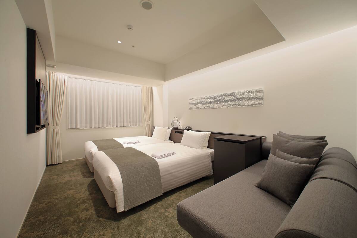 A hotel room with two beds, a sofa, a flat-screen TV, and a window with curtains.