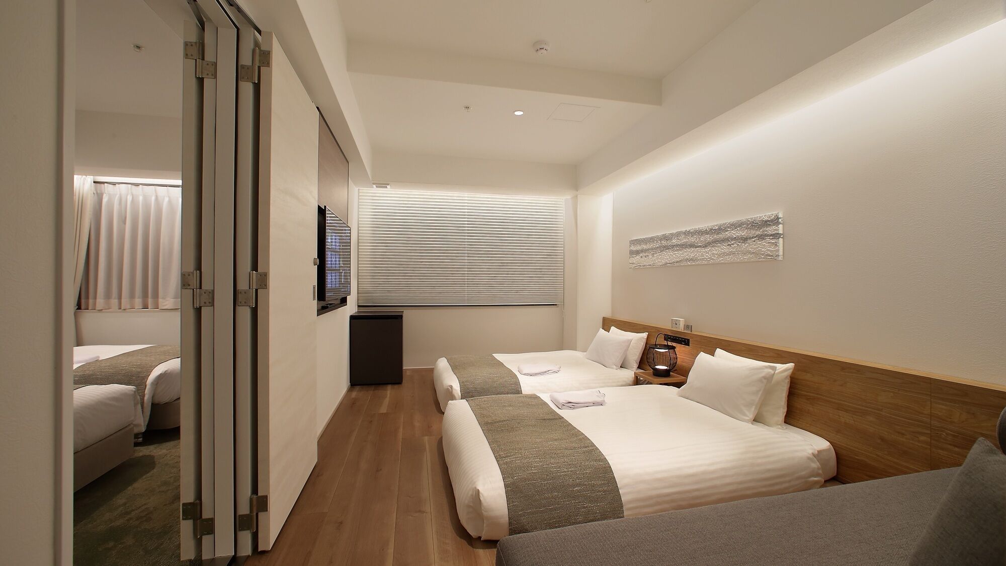 Une chambre d’hôtel avec deux lits, une télévision, une table de chevet avec une lampe et une fenêtre avec des stores.