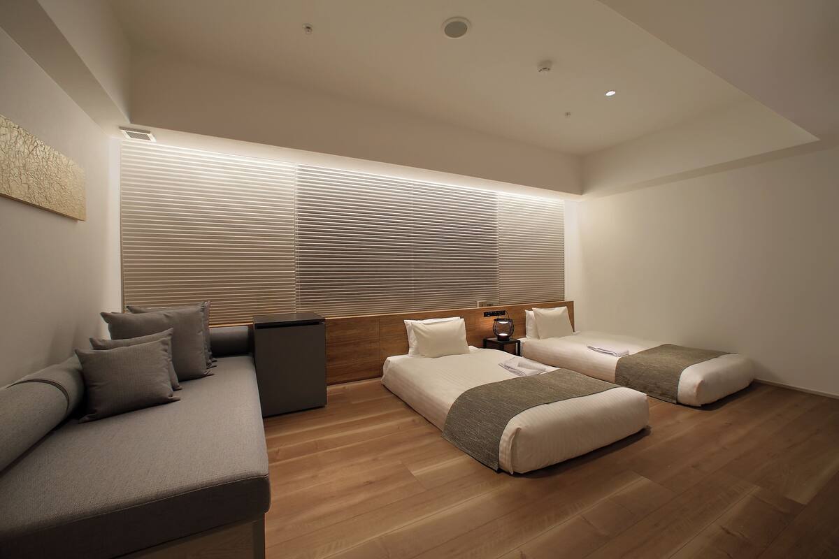 A modern bedroom with a sofa, two beds, wooden flooring, and large windows with blinds.