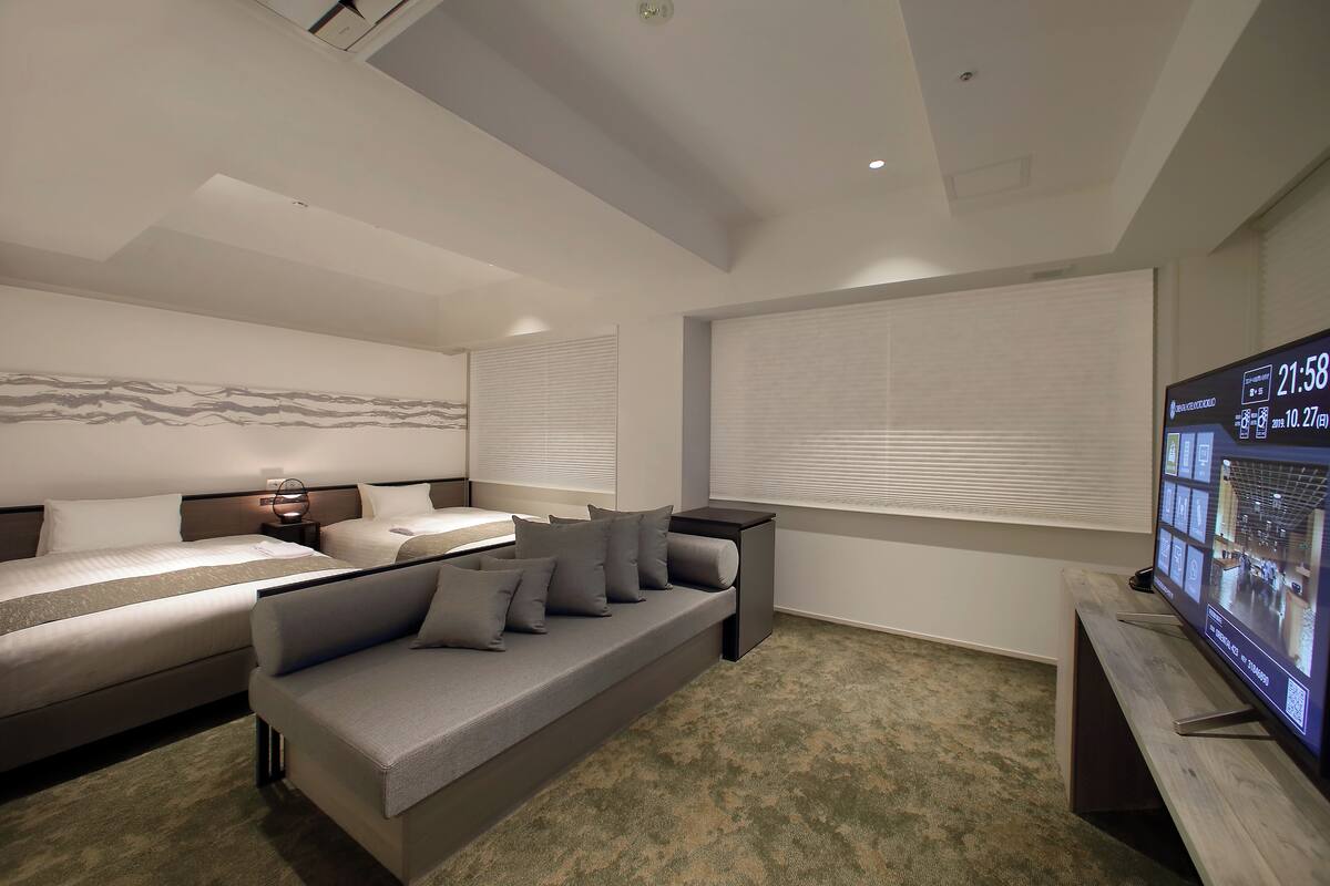 A modern hotel room with a large bed, a sofa, a TV, and a window with blinds.