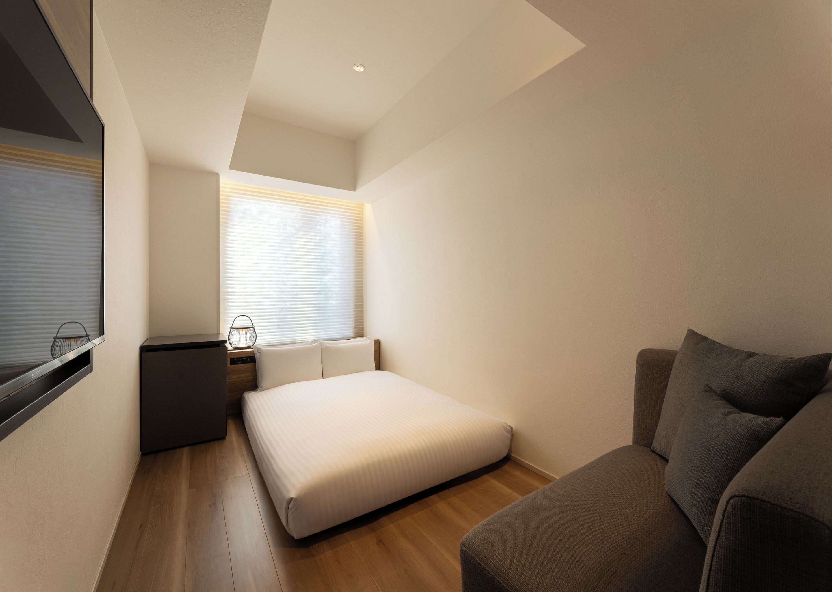A compact bedroom with a single bed, a small bedside table, a chair, and a window with blinds.