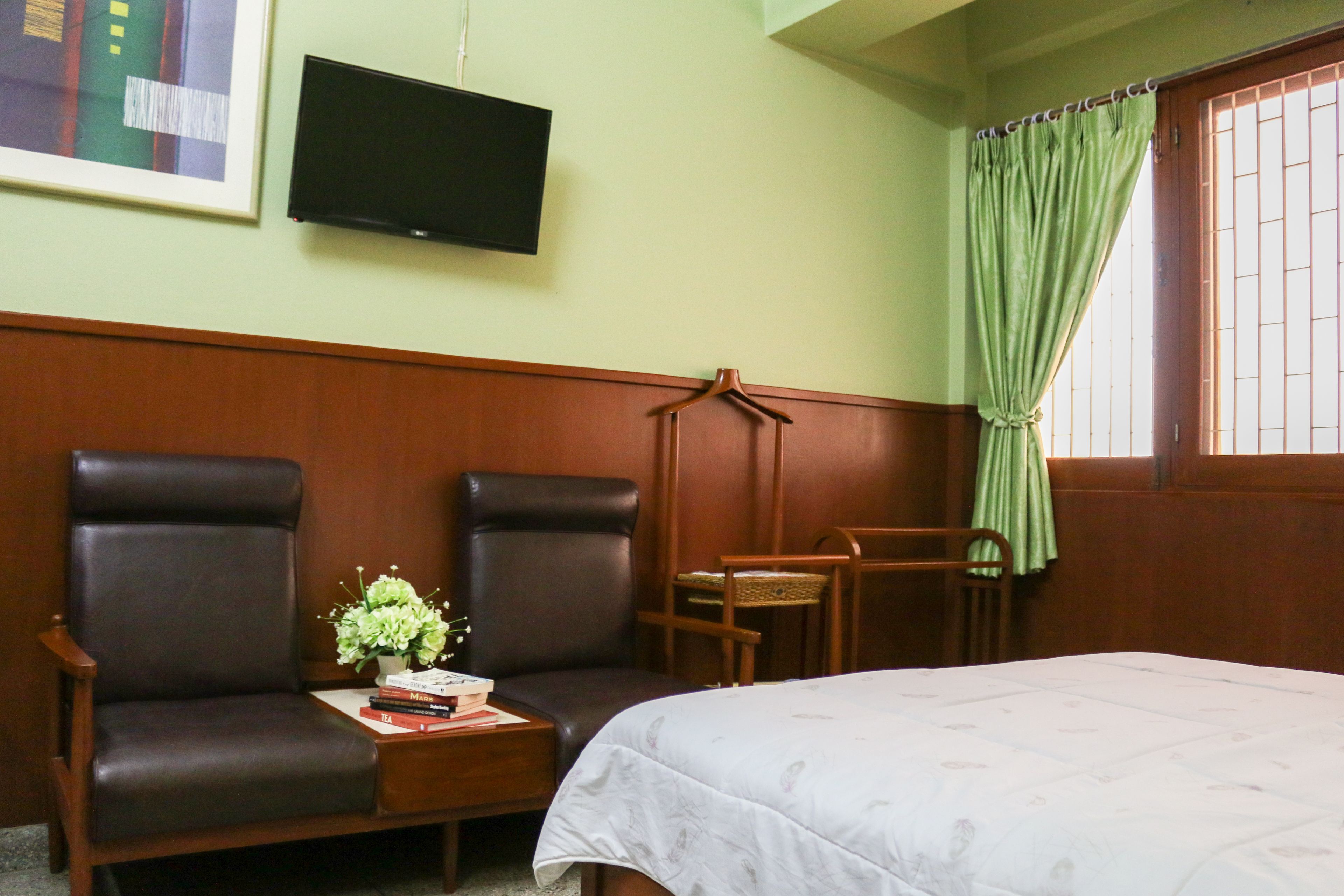 A hotel room with a bed, two armchairs, a small table with a vase of flowers, a television on the wall, and a window with green curtains.