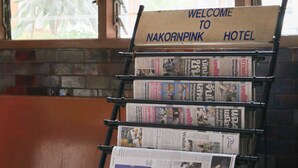 A hotel reception desk with a 'Welcome to Nakornpink Hotel' sign, a newspaper rack with various papers, and a wooden bench.
