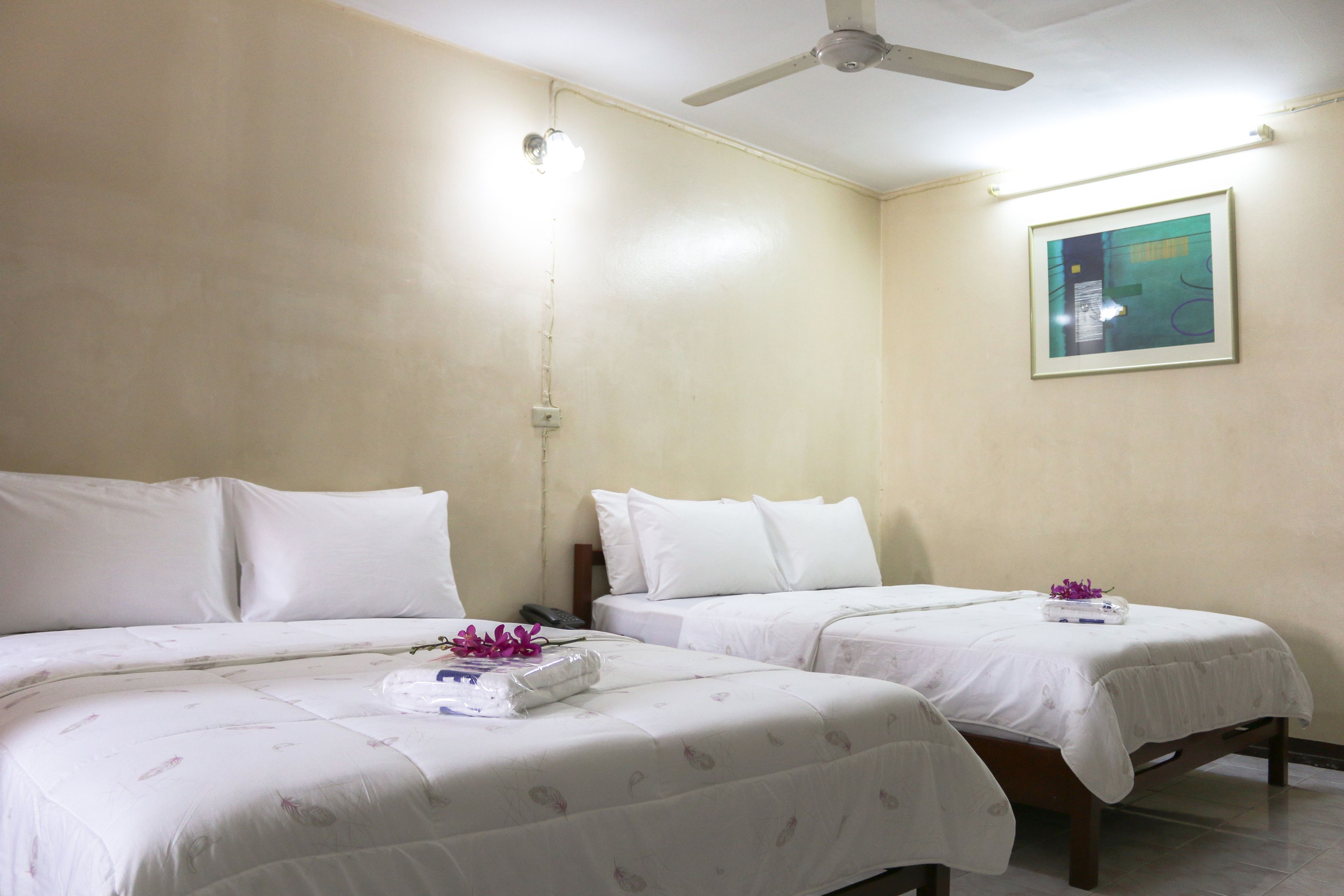 A hotel room with two beds, a ceiling fan, a framed picture on the wall, and a small table with a flower.