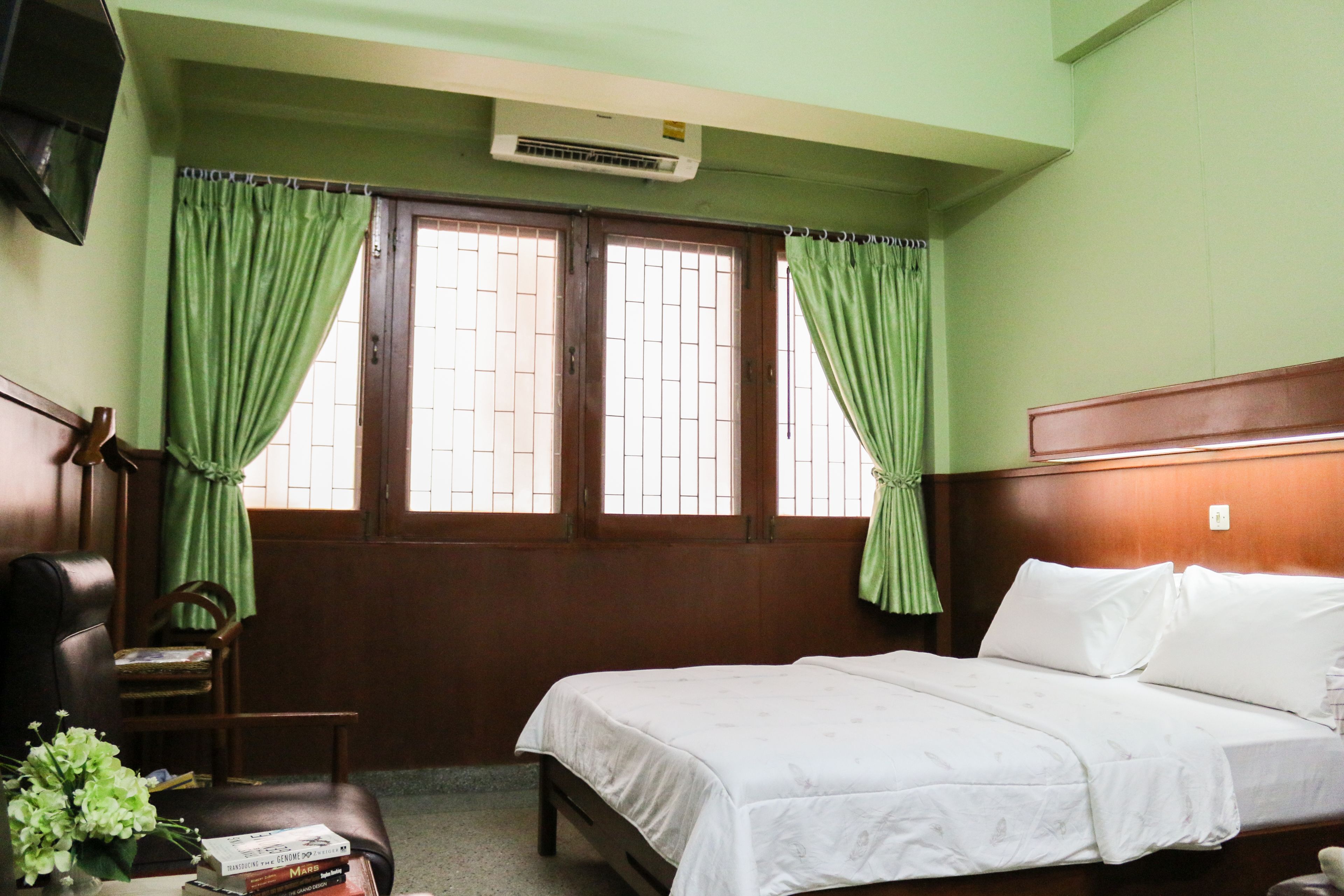 A room with a bed, a chair, a small table with books, and a window with curtains.