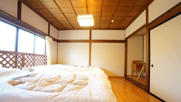 A room with two beds, wooden flooring, a sliding door, and a view through a window with a curtain.