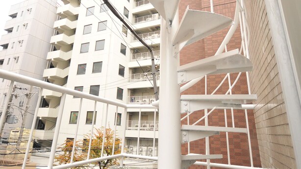 Un escalier en colimaçon avec une rampe blanche, entouré d’une base en béton, dans un environnement urbain avec des bâtiments en arrière-plan.