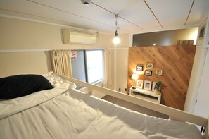 A bedroom with a bed, a window with curtains, a wall-mounted air conditioner, and a bedside table with a lamp and framed pictures. - Condominium House in Osaka Nakatsu (Osaka)