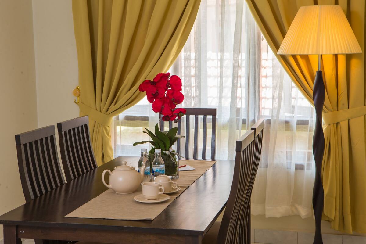 Une table à manger dressée avec une théière, des tasses, des soucoupes et un vase rempli de fleurs rouges.