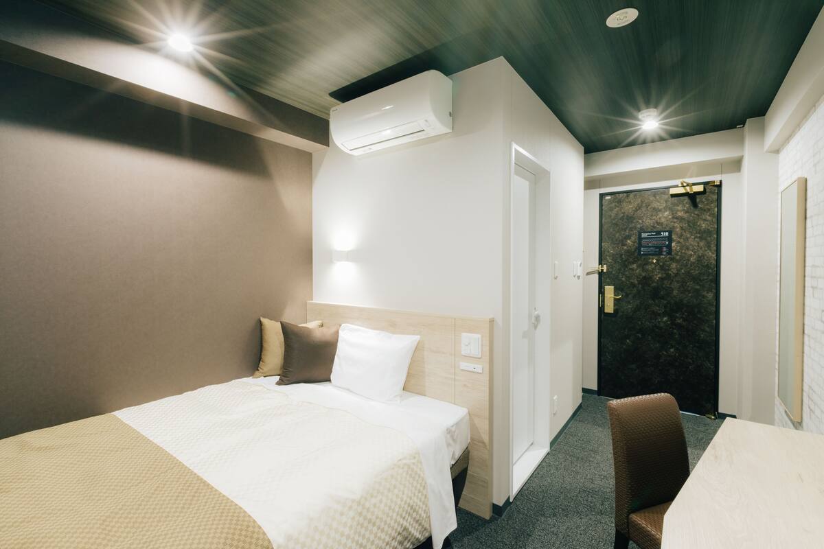A hotel room with a bed, a chair, an air conditioning unit, and a door with a sign.