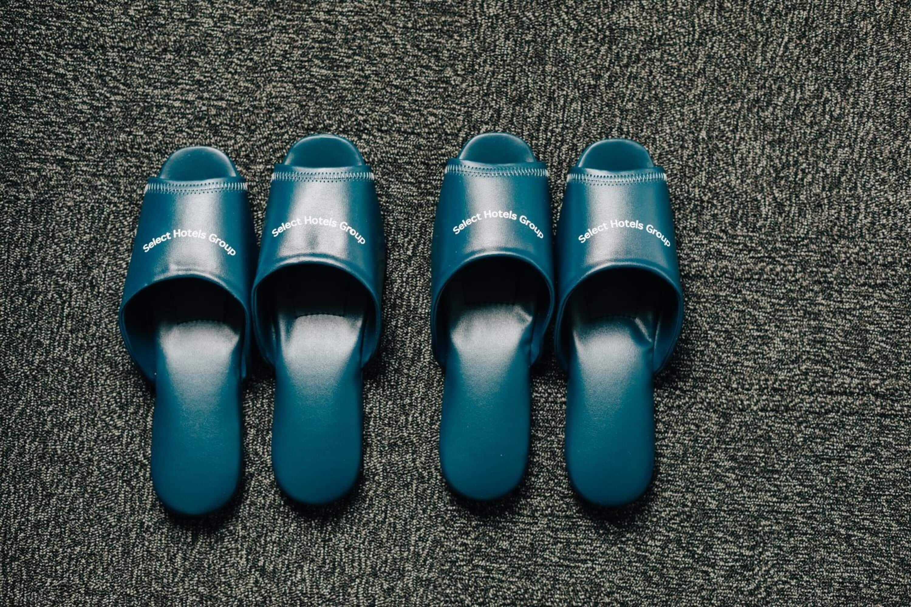 Four pairs of teal slippers with the text "Select Hotels Group" on them, placed on a textured carpet.