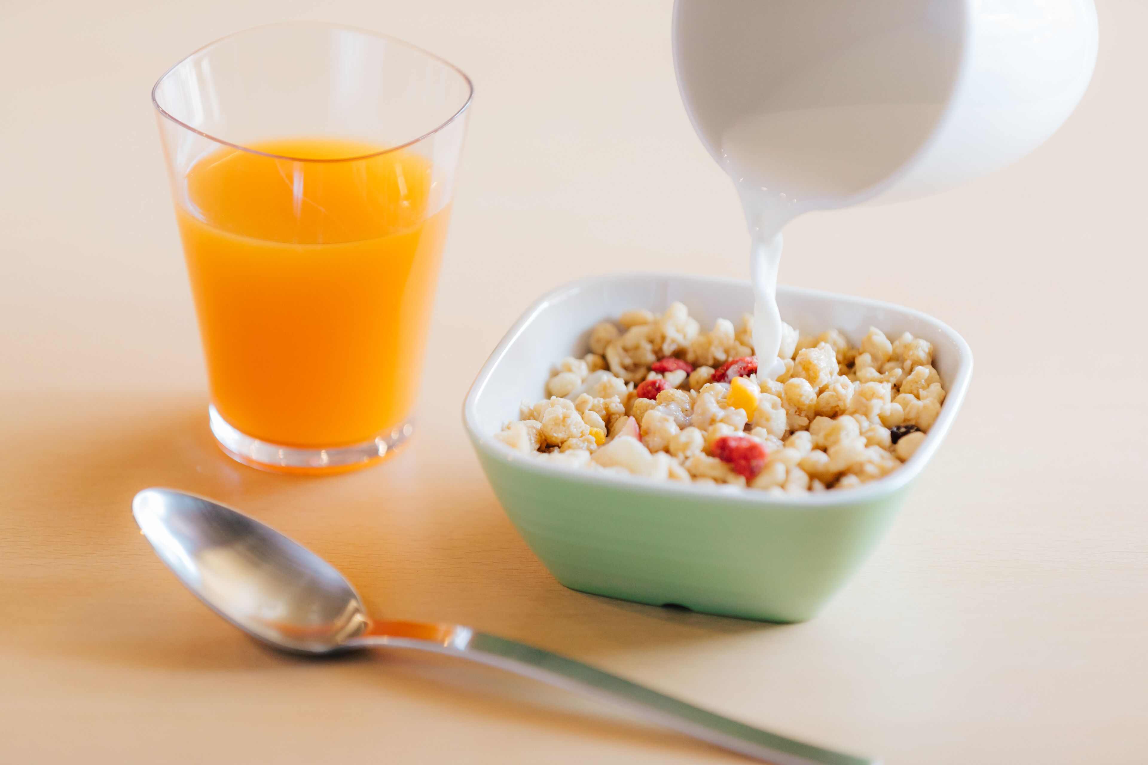 A bowl of cereal with milk being poured into it, next to a glass of orange juice.