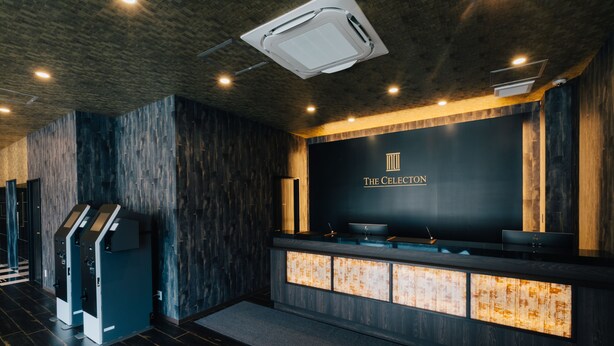 A modern reception area with a dark wood panel wall, a reception desk with a glass top, and a logo above the desk. - The Celecton Kurashiki Mizushima (Kurashiki)