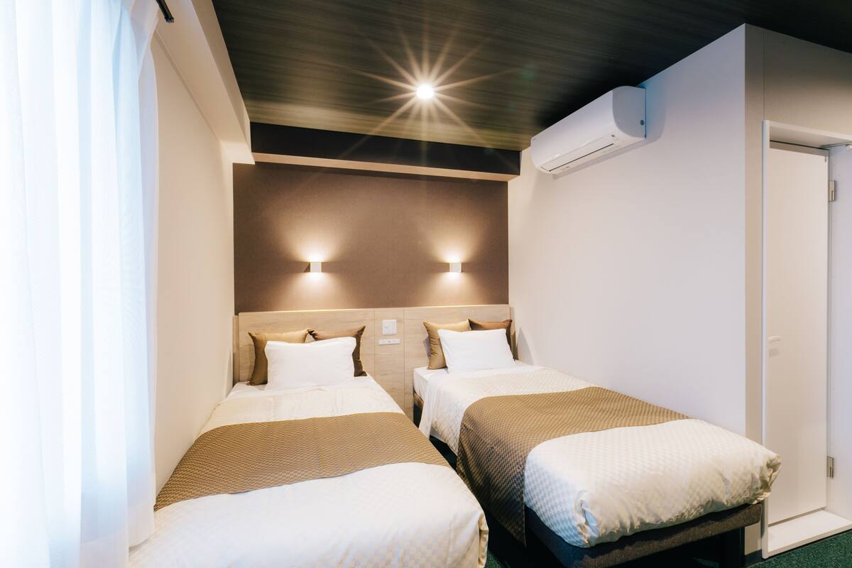 A hotel room with two single beds, a wooden headboard, wall-mounted lights, and an air conditioning unit.