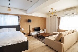 A modern hotel room with a large bed, a sofa, a coffee table, a TV, and an air conditioning unit.