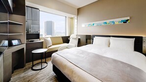 Una habitación de hotel moderna con una cama grande, una zona de estar con vistas, un escritorio con computadora y un ventanal amplio.
