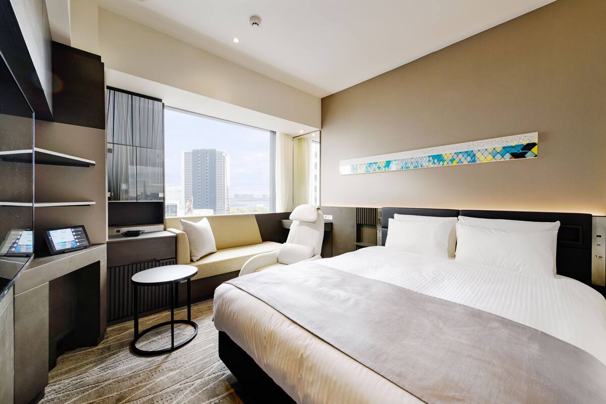 Una habitación de hotel moderna con una cama grande, una zona de estar con vistas, un escritorio con computadora y un ventanal amplio.