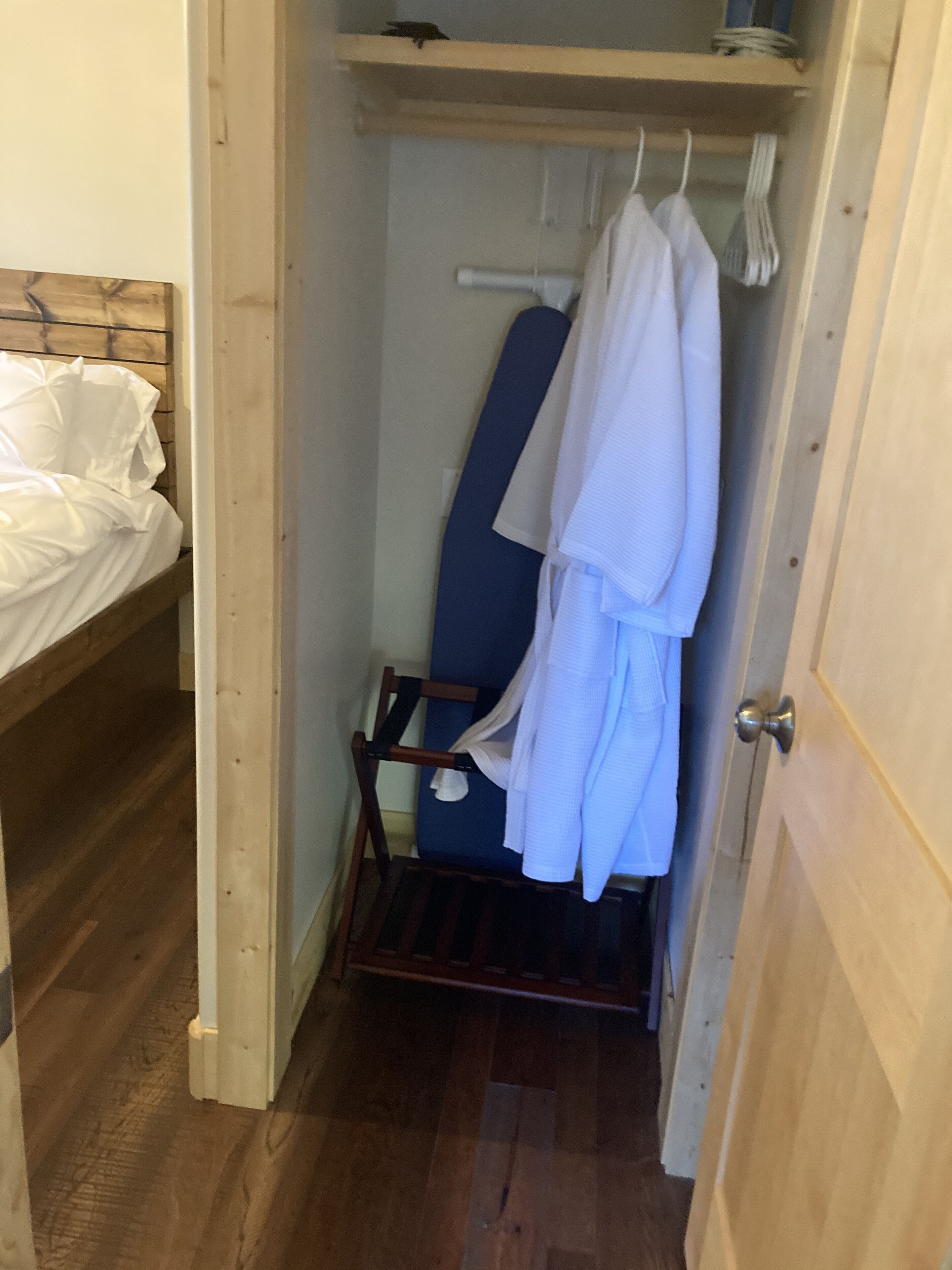 A narrow room with a wooden bed, a small chair, and a hanging rack with clothes.