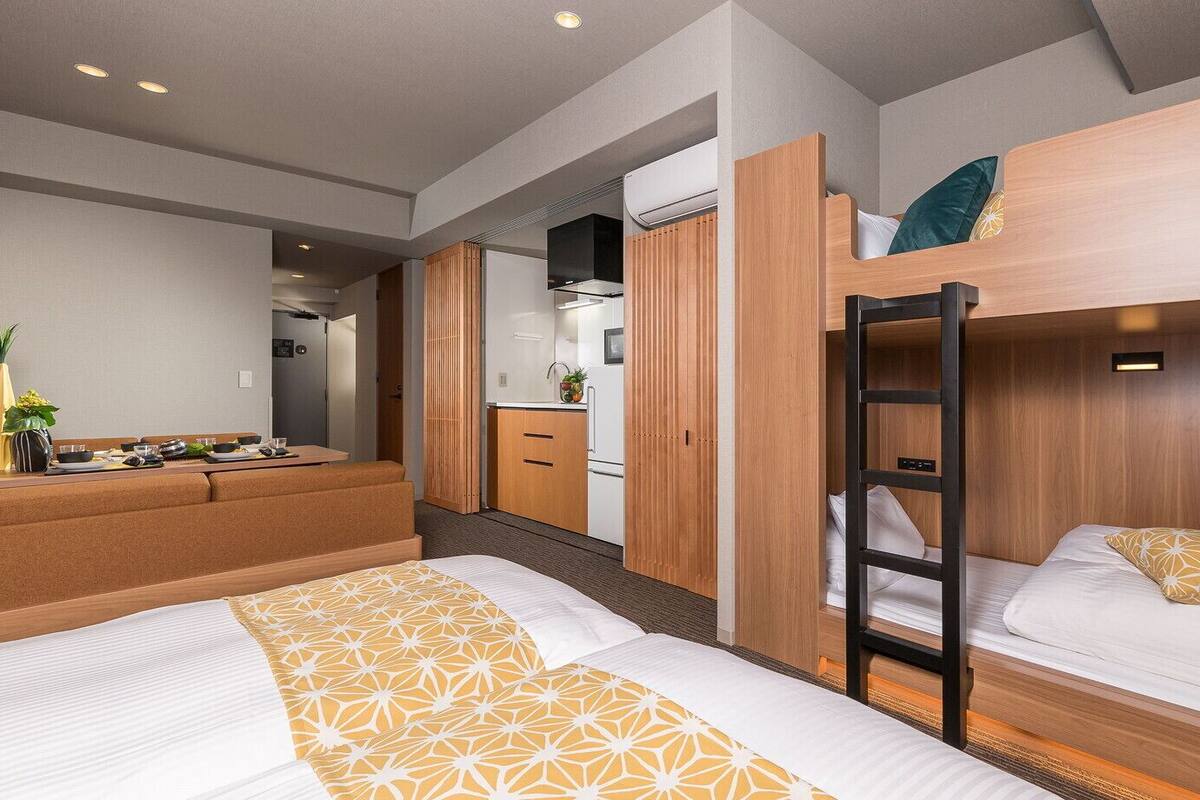 Une chambre d’hôtel moderne équipée de lits superposés, d’une kitchenette et d’un coin salon agrémenté d’une plante.