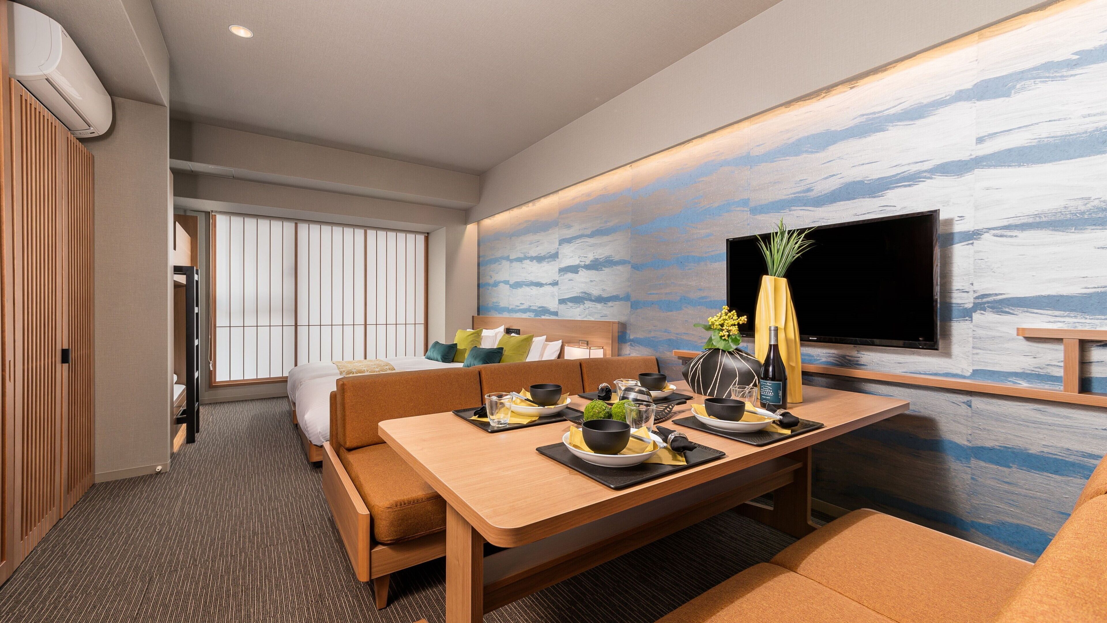 Una habitación de hotel moderna con zona de comedor, sofá, televisor y un ventanal amplio.