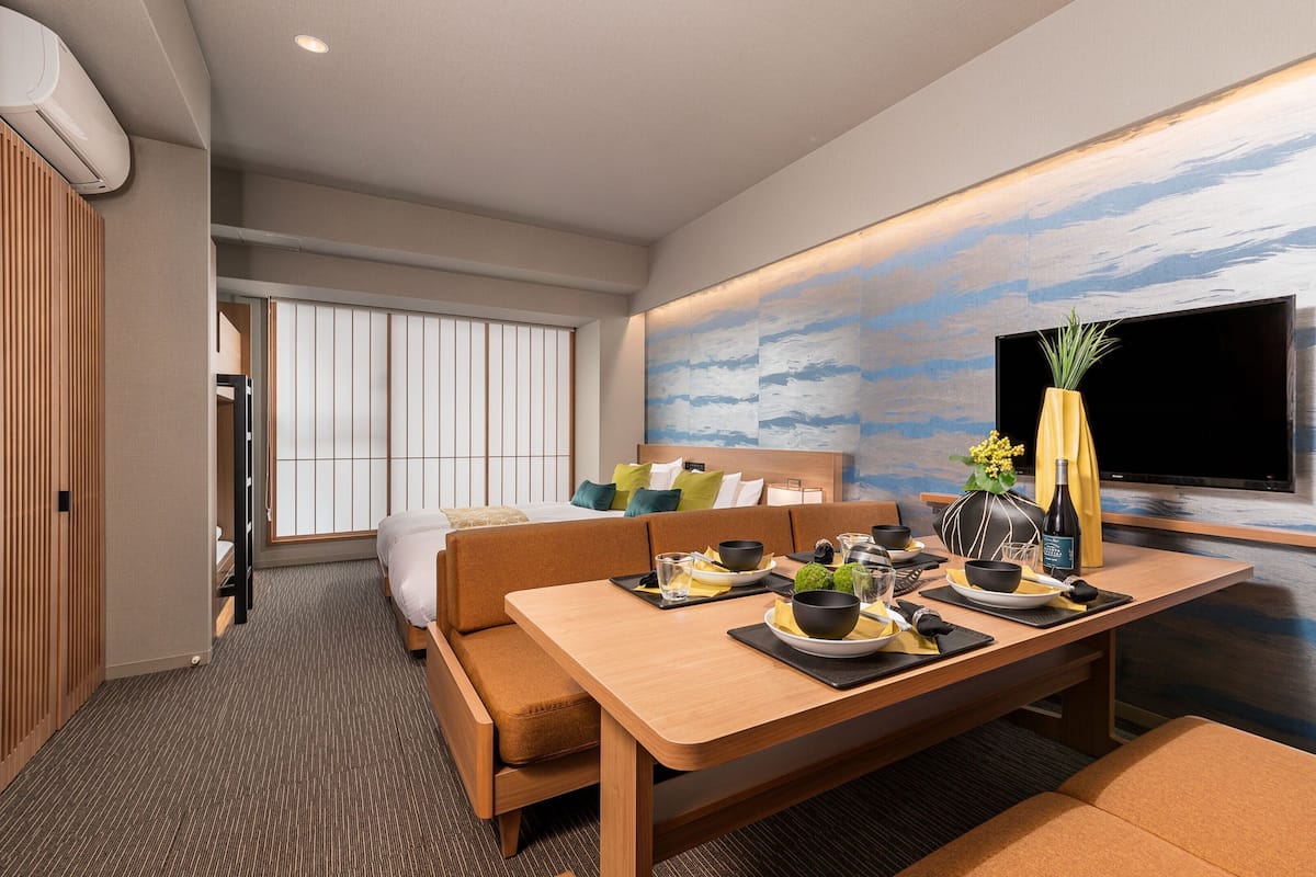 A modern hotel room with a dining area, a sofa, a TV, and a large window.