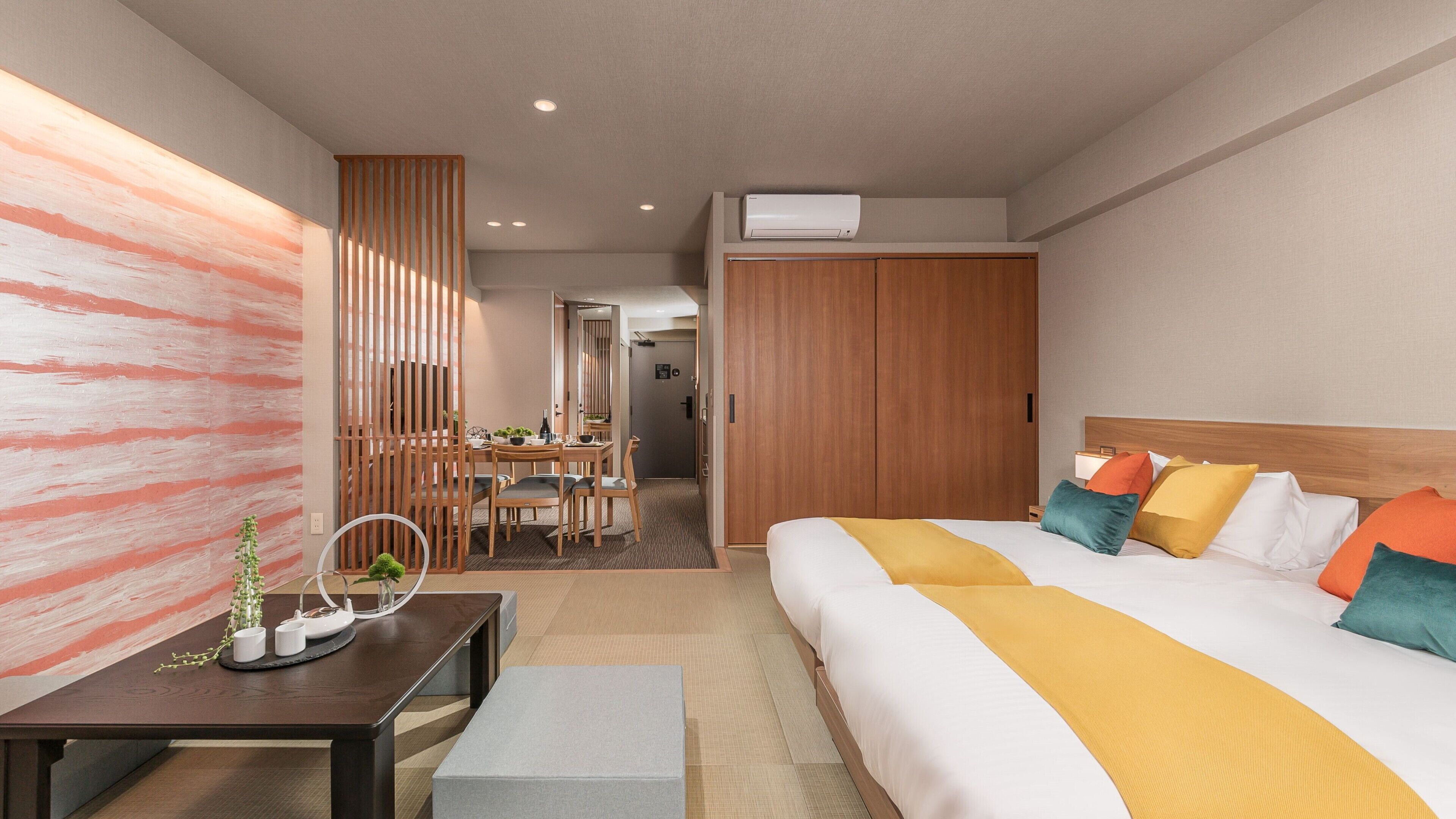 Habitación de hotel con cama, mesita de noche y escritorio con silla. Se observa un armario de madera y una puerta que da a otra habitación.