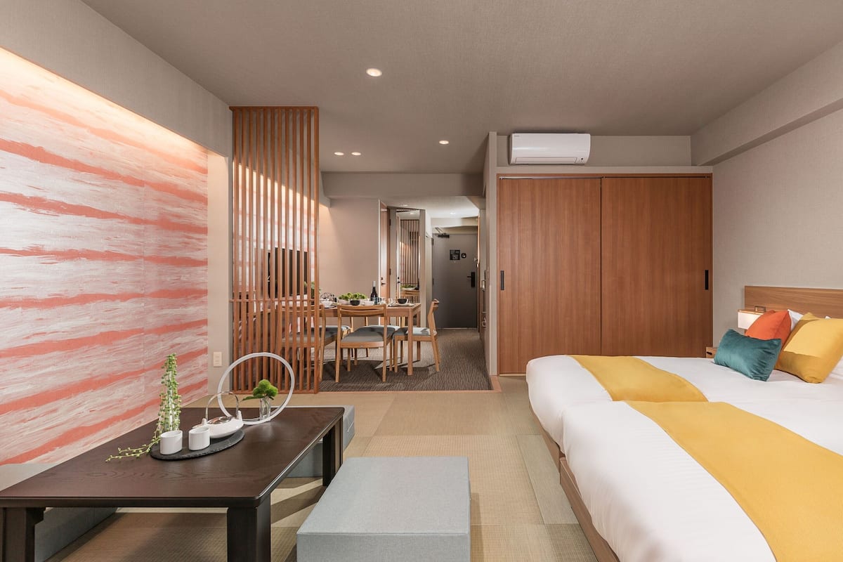 A hotel room with a bed, bedside table, and a desk with a chair. There is a wooden wardrobe and a door leading to another room.