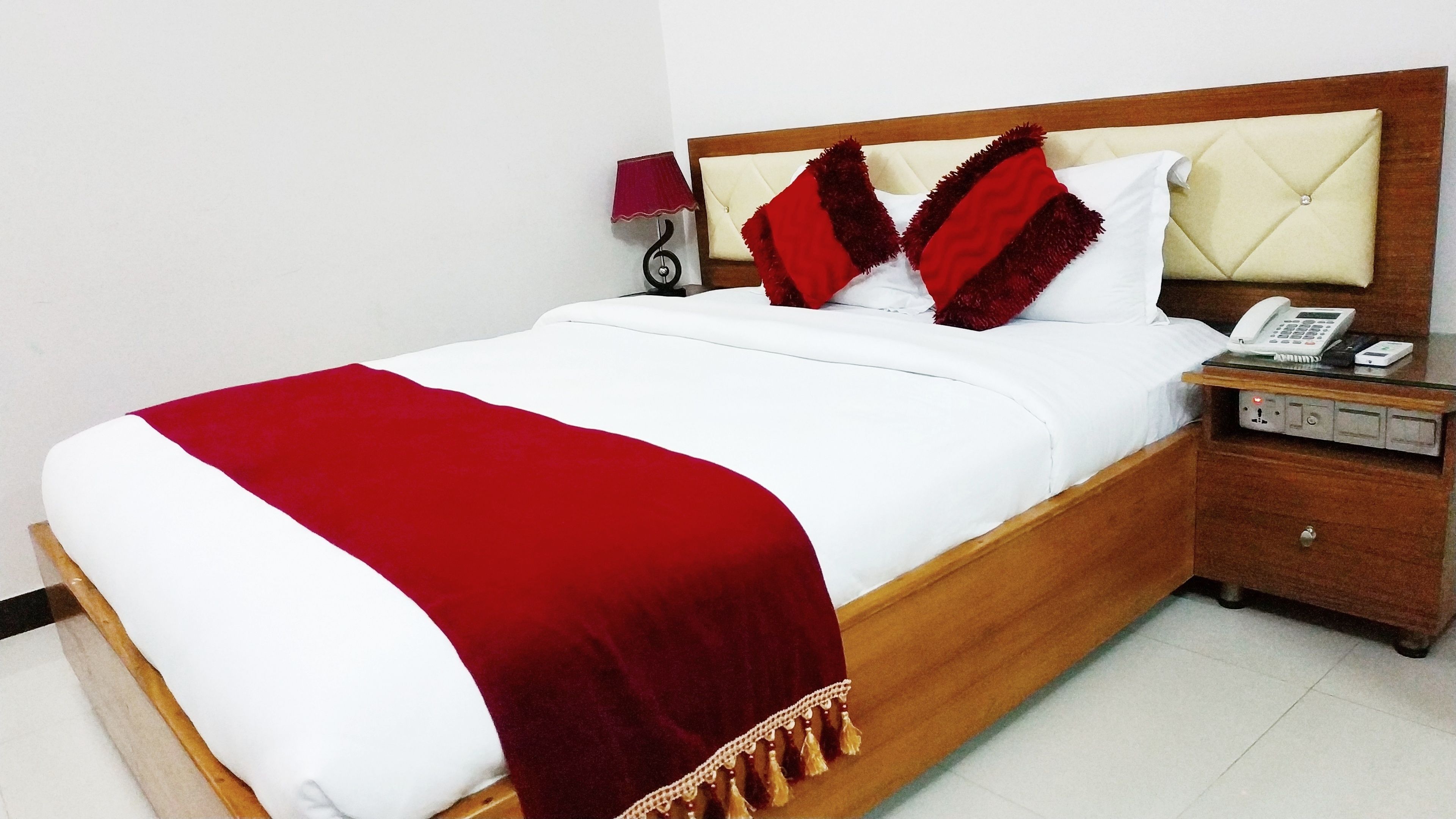 A hotel room with a wooden bed, white bedding, a red throw blanket, a bedside table with a phone, and a lamp.
