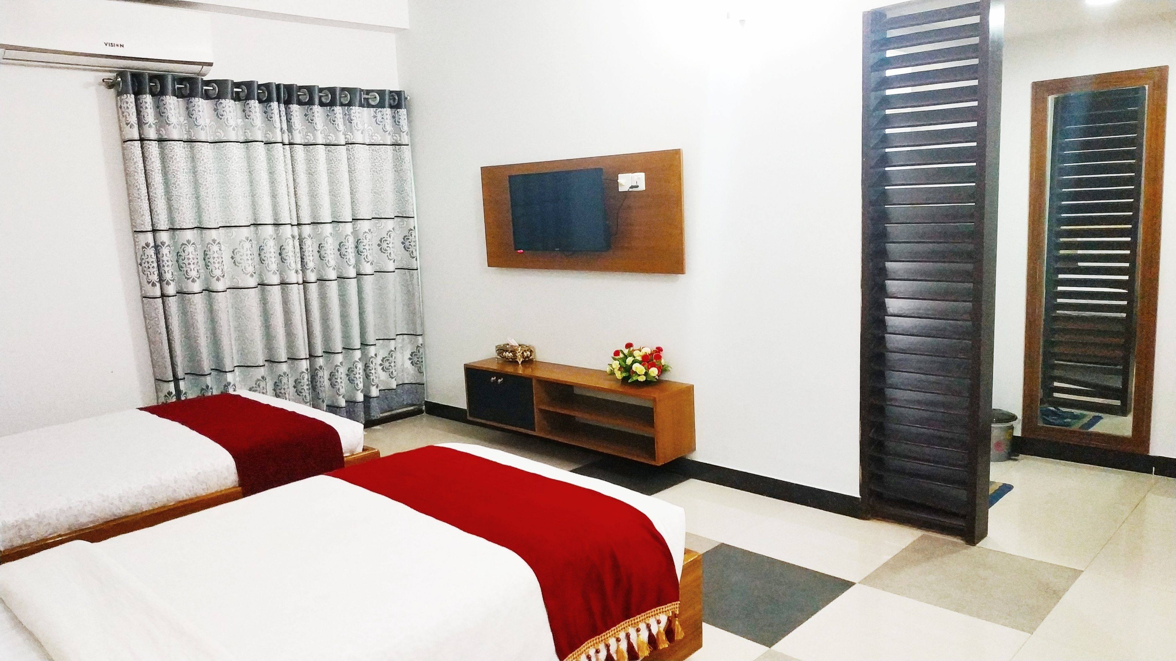 A hotel room with two beds, a wooden console table with a vase of flowers, a wall-mounted TV, and a sliding door.