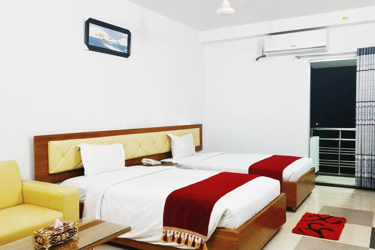 A hotel room with two beds, a yellow armchair, a bedside table with a tissue box, and a red rug.