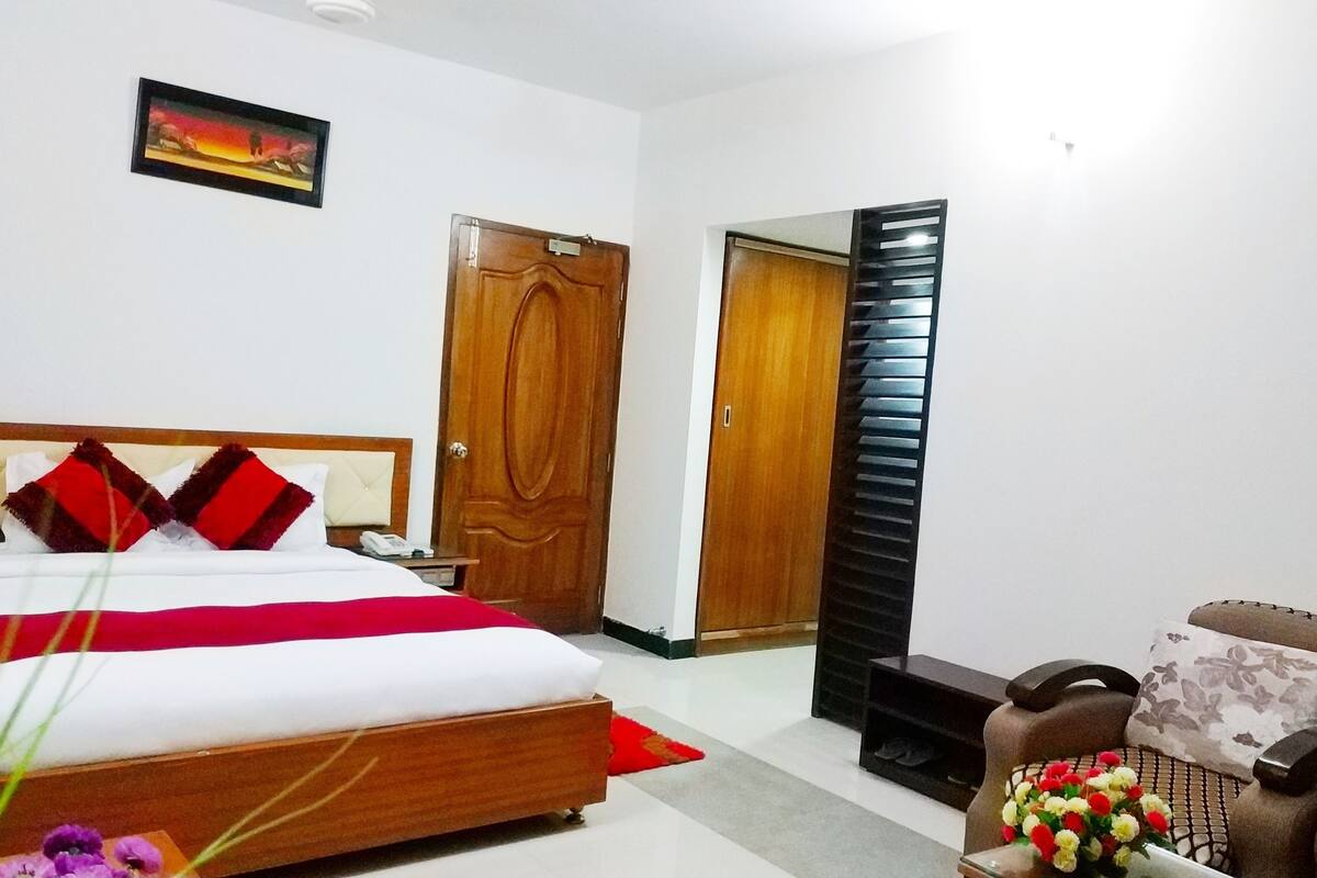 A hotel room with a bed, a wooden door, a small table with a vase of flowers, and a chair.