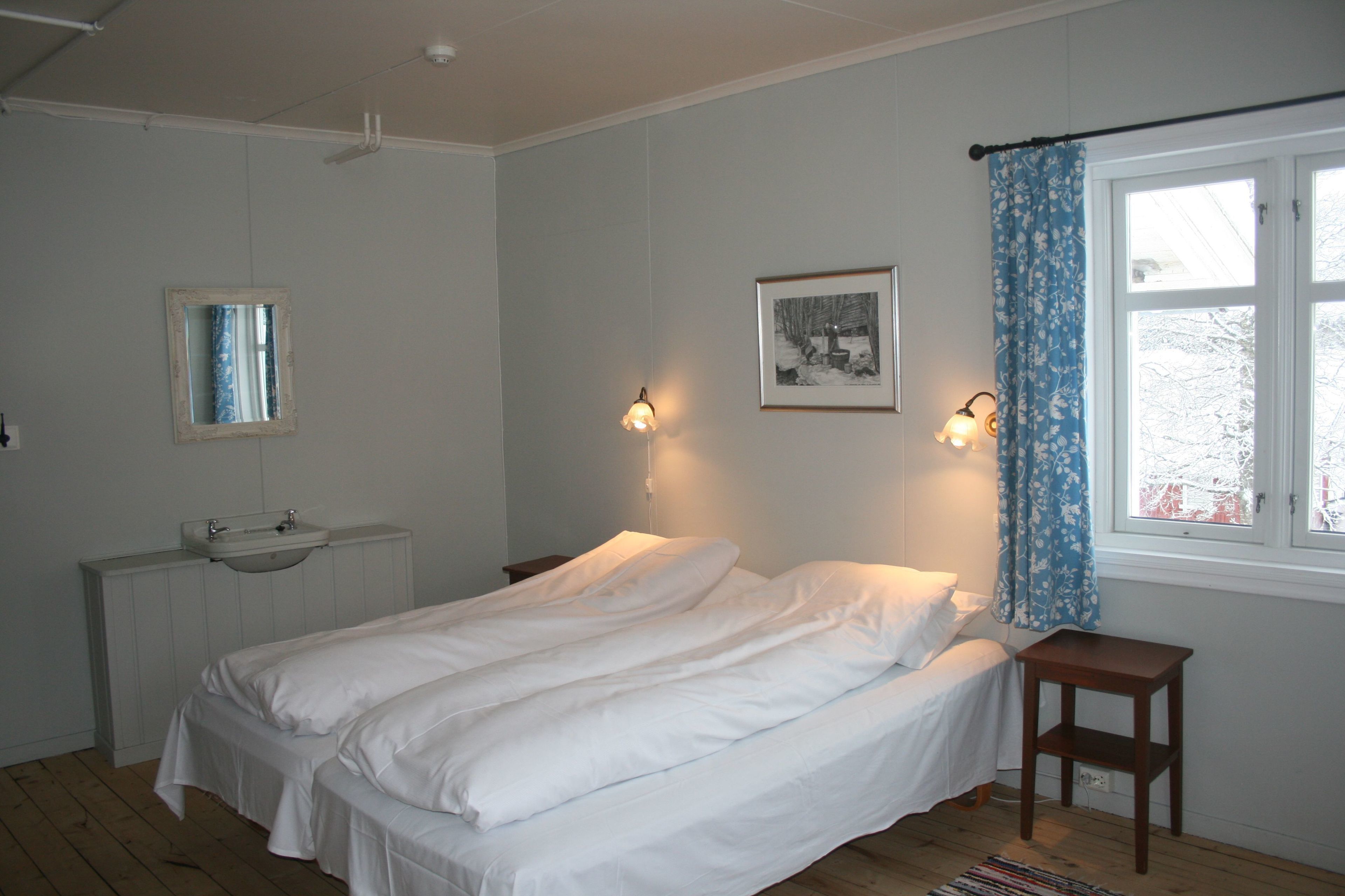 Une chambre à coucher comprenant un lit, une table de chevet, une fenêtre avec des rideaux, un luminaire fixé au mur et un tableau encadré.