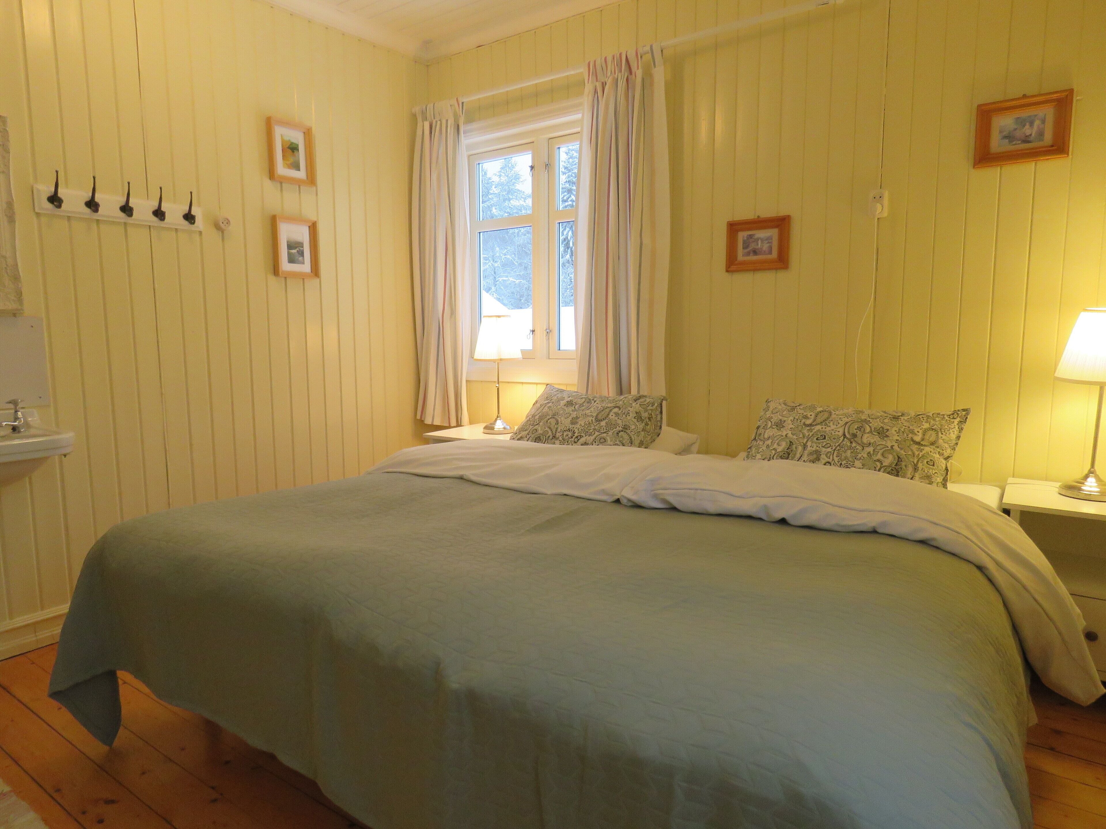 A bedroom with a bed, a bedside table with a lamp, a window with curtains, a wall-mounted shelf with hooks, and a small picture frame.