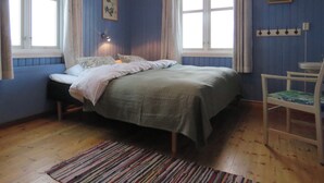 Quarto com cama, duas janelas com cortinas, mesa de cabeceira, cadeira, tapete e prateleira fixada na parede.