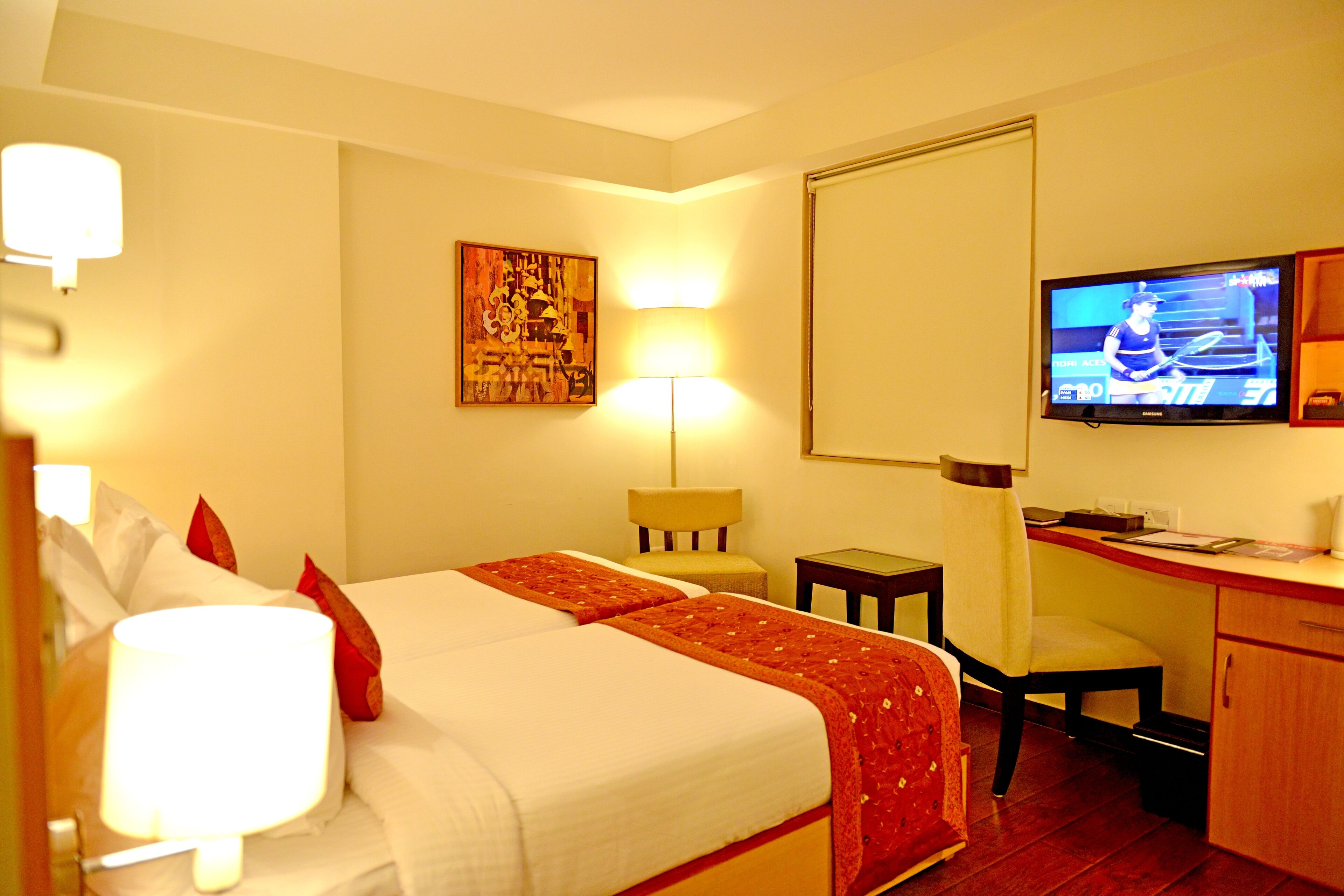A hotel room with two beds, a desk, a chair, a television, and a lamp.