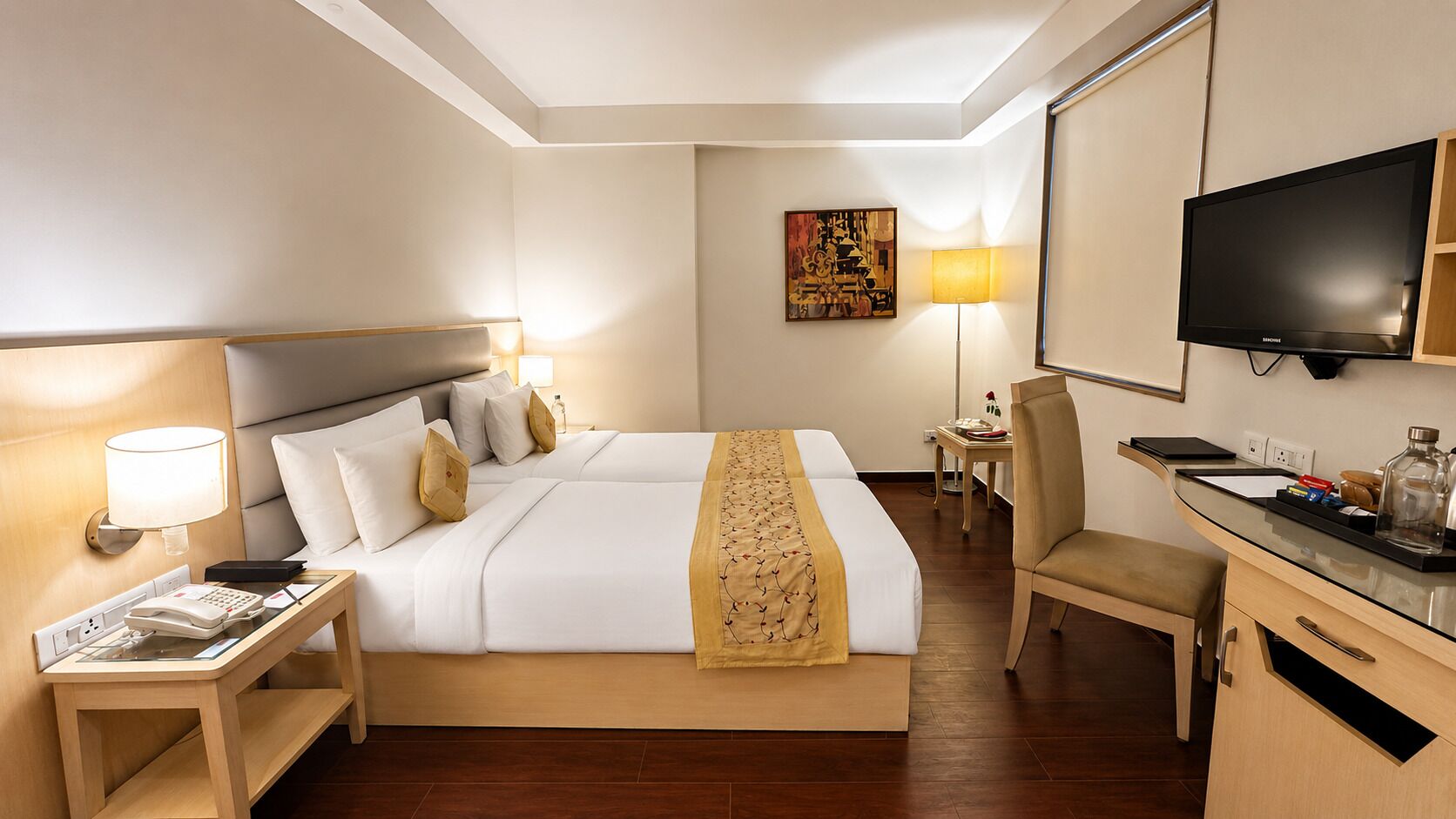 A hotel room with a bed, bedside table, lamp, desk, chair, TV, and artwork on the wall.