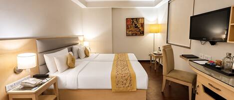 Habitación de hotel con cama, mesita de noche, lámpara, escritorio, silla, televisor y una obra de arte en la pared.