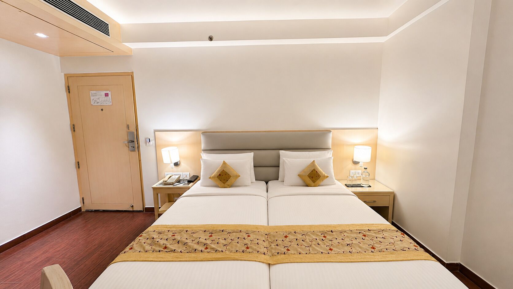 A hotel room with a double bed, two bedside tables with lamps, a wooden door, and a patterned bedspread.