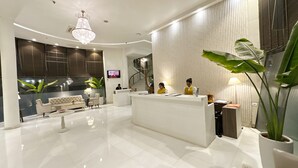 A hotel reception area with a white reception desk, a sofa, a chandelier, and a television on the wall. - The Theme Hotel Jaipur (Jaipur)