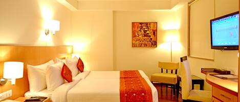 Quarto de hotel com cama, mesa de cabeceira, luminária, escrivaninha, cadeira, televisão e obra de arte na parede.