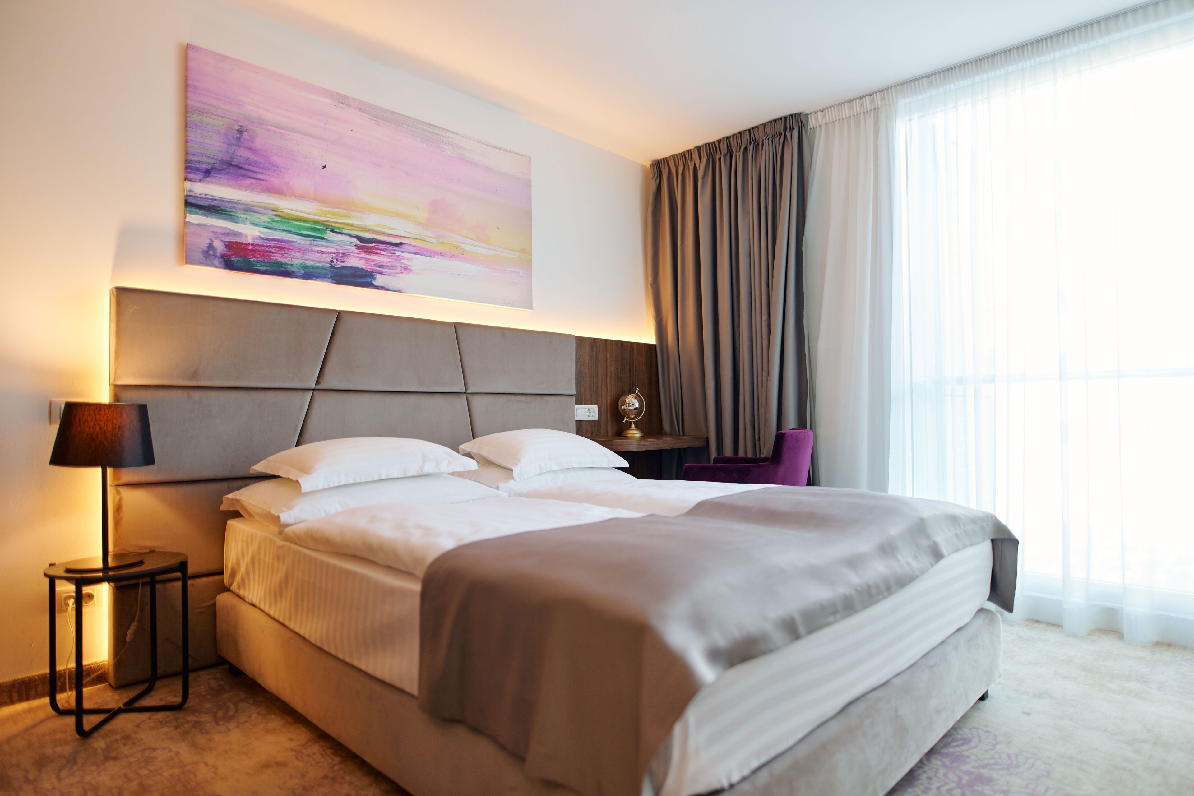 A modern hotel room with a large bed, a bedside table with a lamp, a nightstand, a chair, and a large window with curtains.