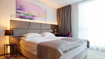 Ein modernes Hotelzimmer mit einem großen Bett, einem Nachttisch mit Lampe, einem Sessel und einem großen Fenster mit Vorhängen.
