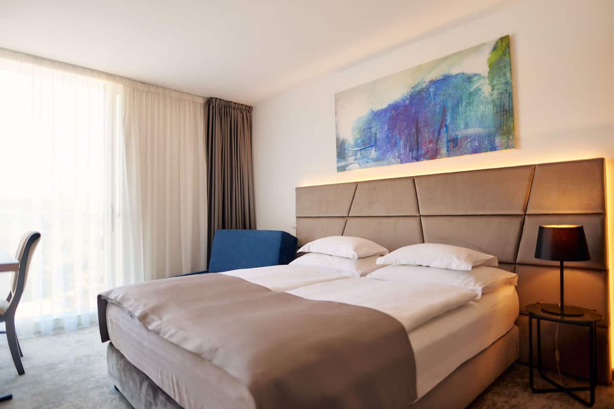 A hotel room with a large bed, a desk, and a chair. There is a painting on the wall and a lamp on the side table.